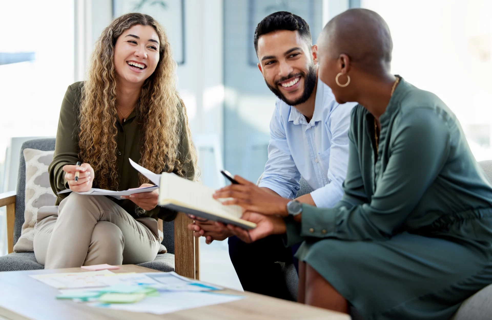 Three people sit together, smiling and talking while holding notebooks and papers, suggesting a collaborative meeting or discussion about health plan compliance risk in a bright, modern office setting.