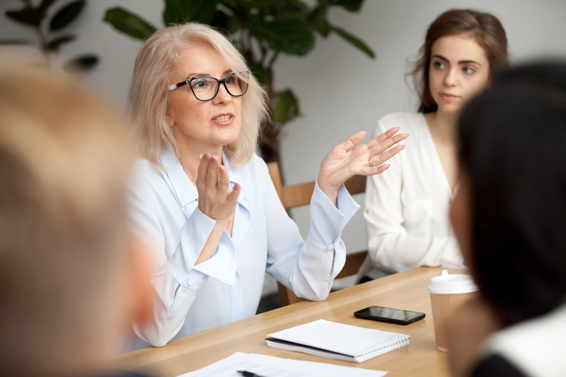 A woman with blonde hair and glasses speaks animatedly during a meeting about managing risk, with notebooks, a smartphone, and a coffee cup in front of her. Other people listen attentively in the background.