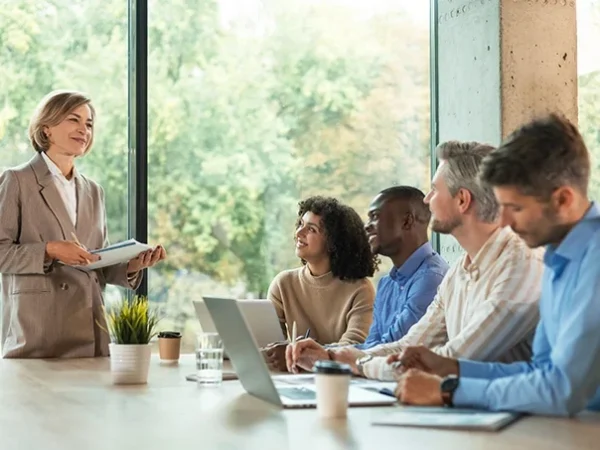 A woman stands presenting to four seated colleagues at a conference table with laptops, notebooks, and coffee cups; large windows show greenery outside as they discuss how better data can provide a strategic advantage.