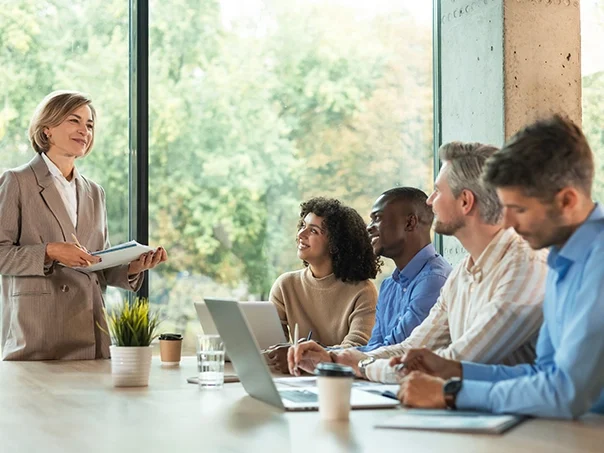A woman stands presenting to four seated colleagues at a conference table with laptops, notebooks, and coffee cups; large windows show greenery outside as they discuss how better data can provide a strategic advantage.