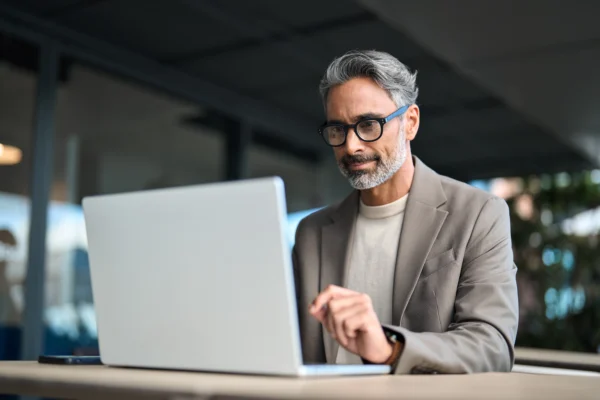 A middle-aged man with gray hair and glasses sits at a desk, working on his laptop. Dressed in a beige blazer, he appears focused as he reviews health plan distribution systems in a modern, outdoor setting.